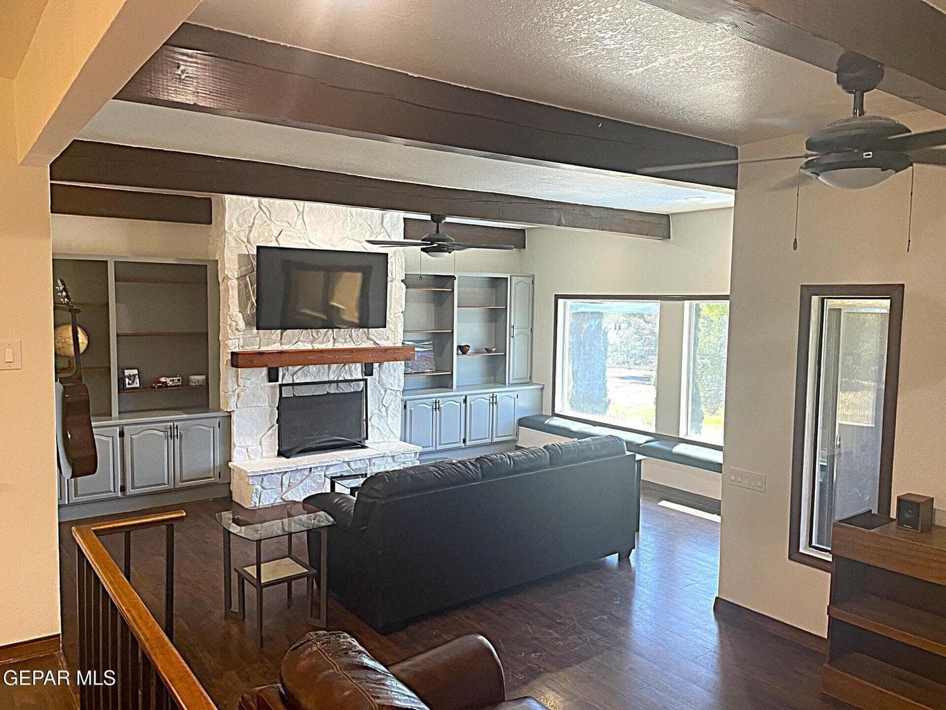 Exposed ceiling beams, stone-faced fireplace with wood mantel, built-in shelving and cabinetry, wood-finish flooring, and large picture windows