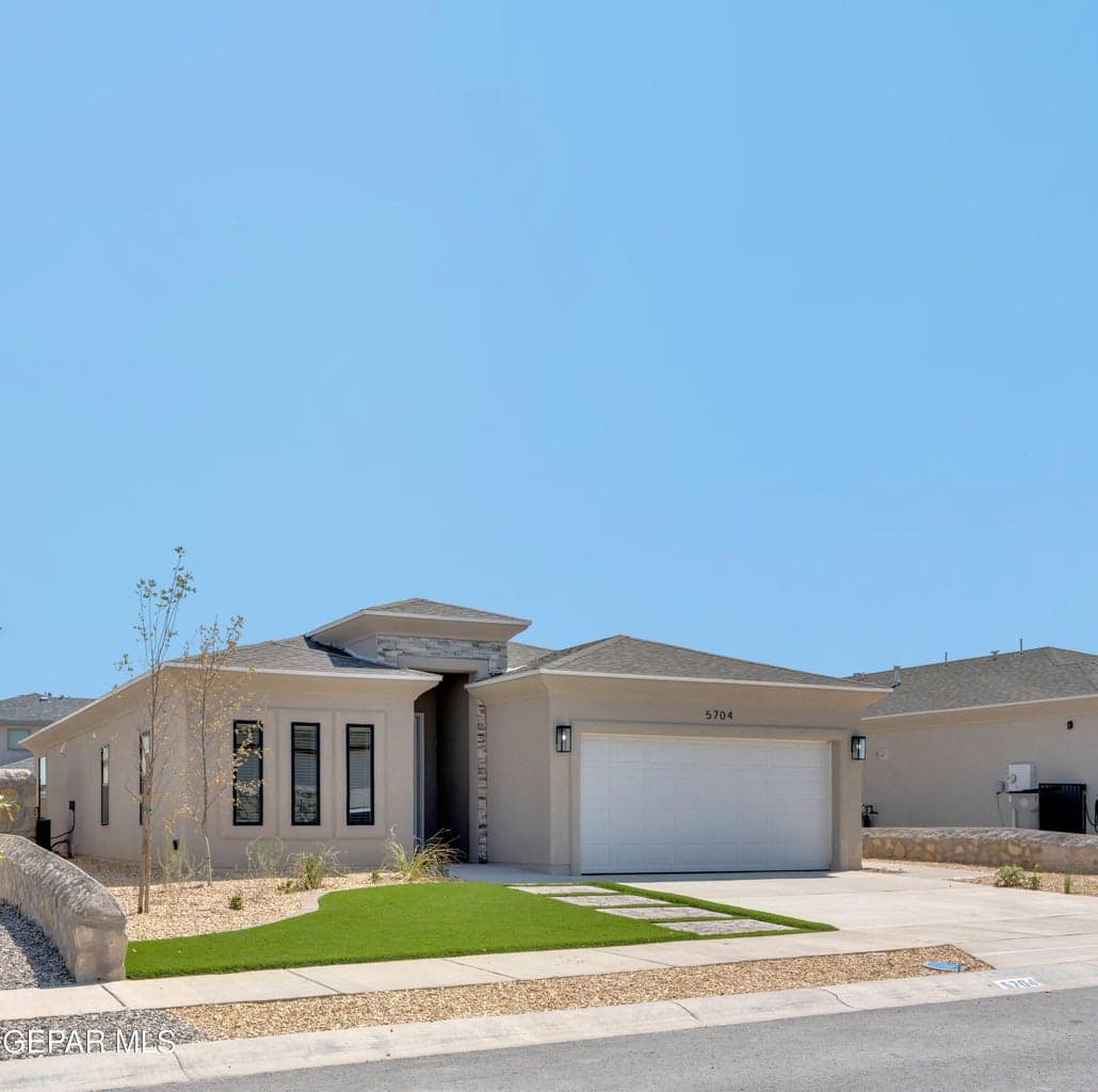 Contemporary stucco exterior with a stone accent entry, attached two-car garage, and a low-maintenance landscape featuring artificial turf and concrete stepping stones