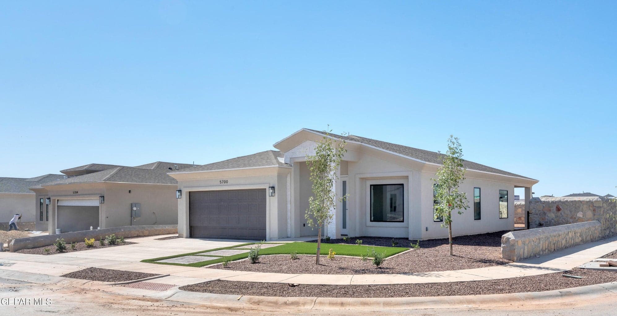 Contemporary stucco exterior, attached two-car garage, low-maintenance landscaping with artificial turf accents, stone retaining wall, and modern exterior lighting