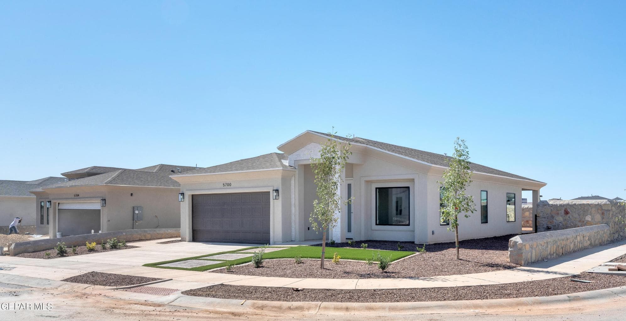 Contemporary stucco exterior, attached two-car garage, low-maintenance landscaping with artificial turf accents, stone retaining wall, and modern exterior lighting