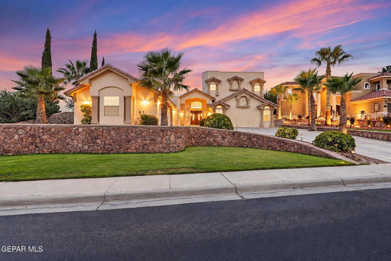 Stucco exterior with multiple rooflines, stone retaining wall, mature palm trees, and a paved driveway