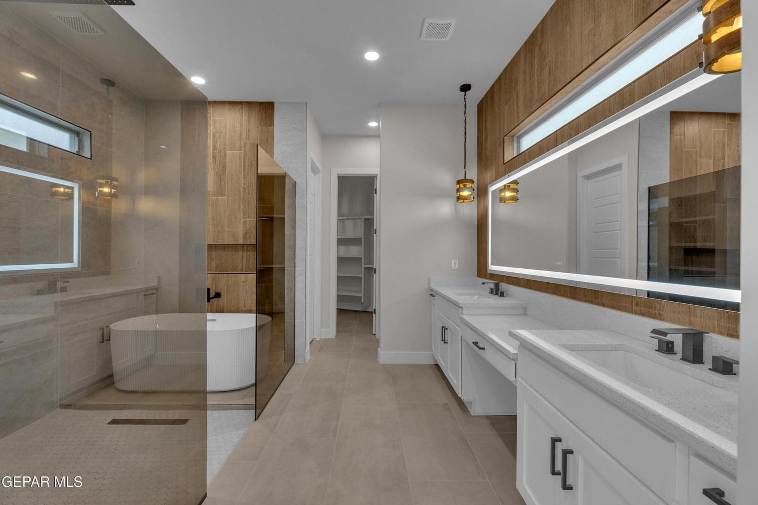 Dual vanity with integrated sinks, back-lit mirror, wood-finish accent wall, freestanding soaking tub, and walk-in shower with glass enclosure