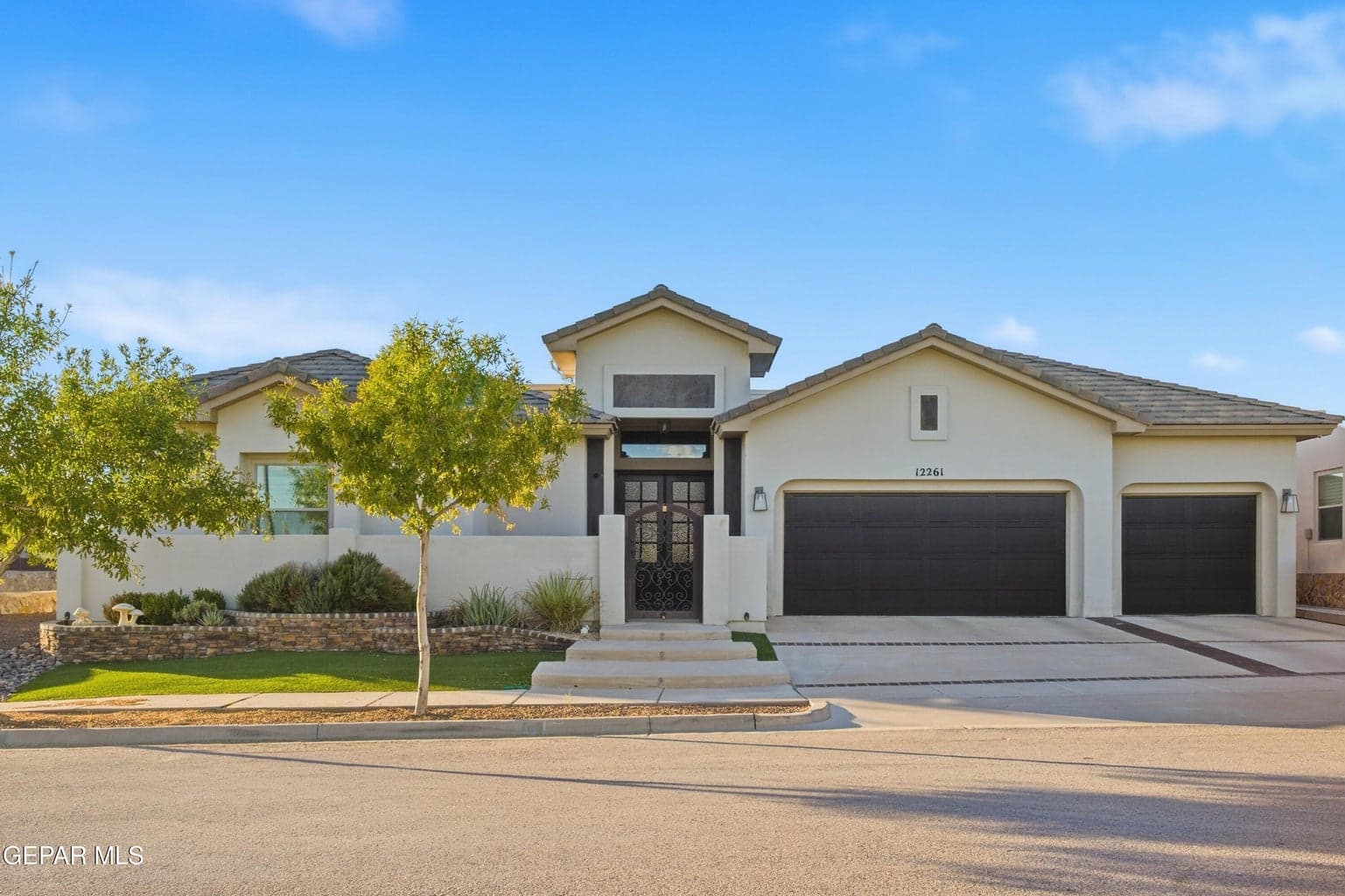 Stucco exterior with a tiled roof, featuring a grand entryway with an iron door, a manicured front lawn, and a three-car garage