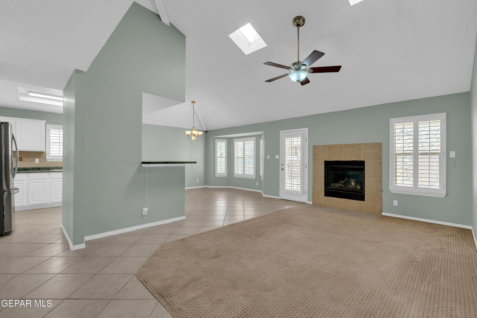 Vaulted ceilings featuring multiple skylights, fireplace with tile surround, tile flooring, ceiling fan with integrated lighting, and plantation-style window shutters