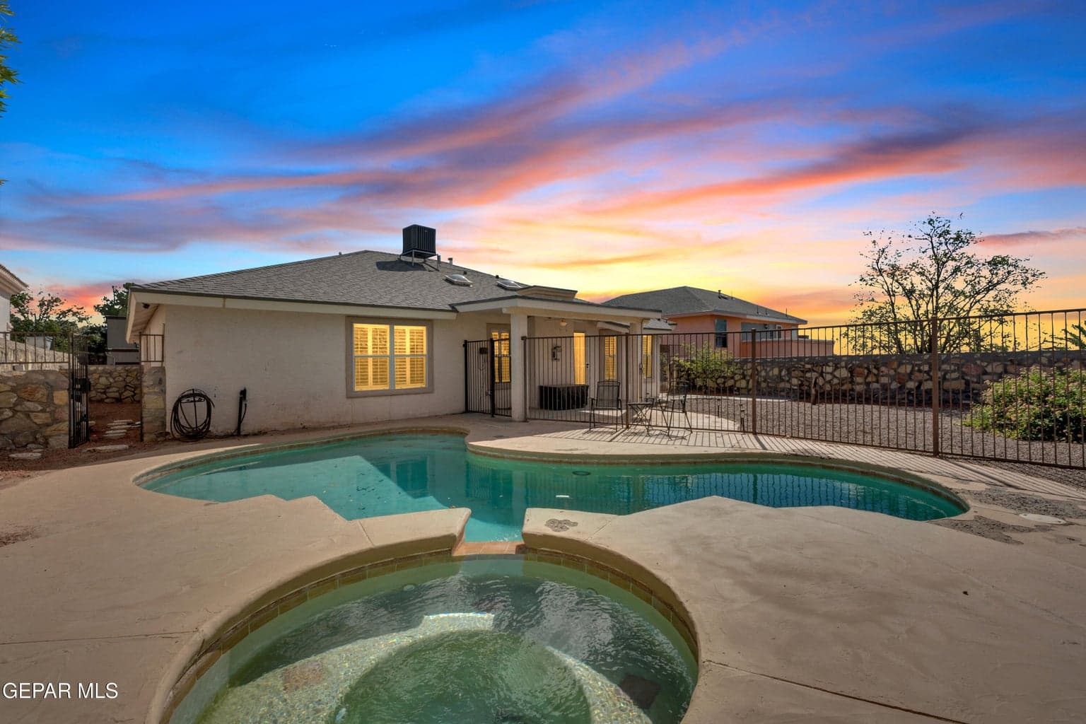 Rear exterior featuring a swimming pool, integrated spa, concrete decking, stone perimeter wall, and metal fencing