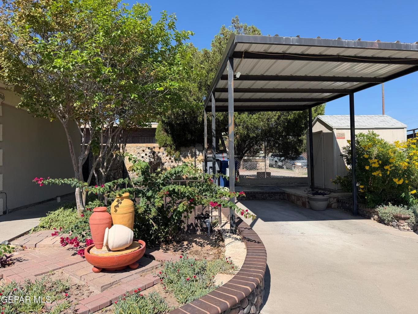 Concrete carport with a metal roof, curved brick retaining wall, and landscaped garden area