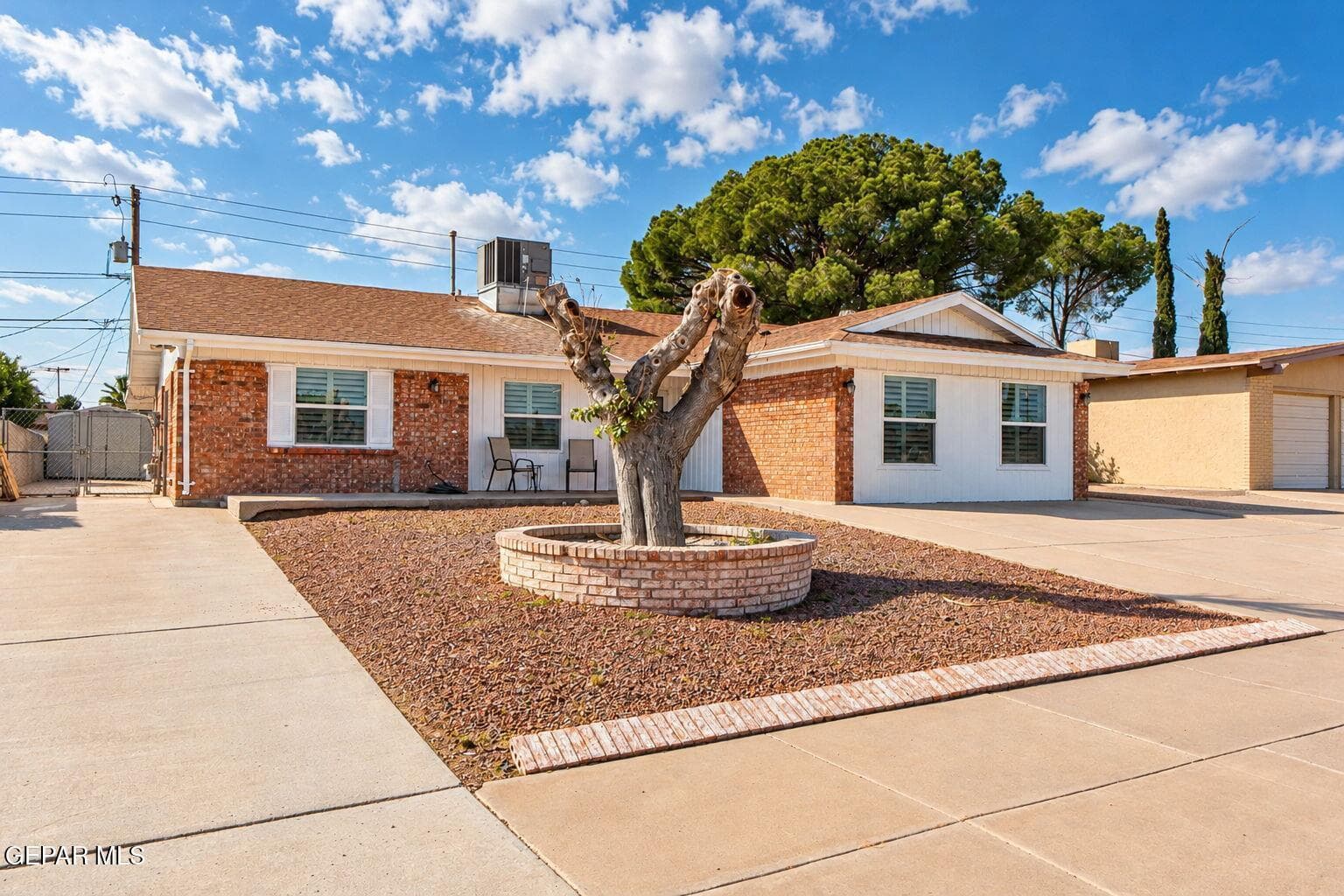 Single-story residence with a brick and stucco exterior, prominent circular brick landscape planter, expansive concrete driveway, and white window shutters