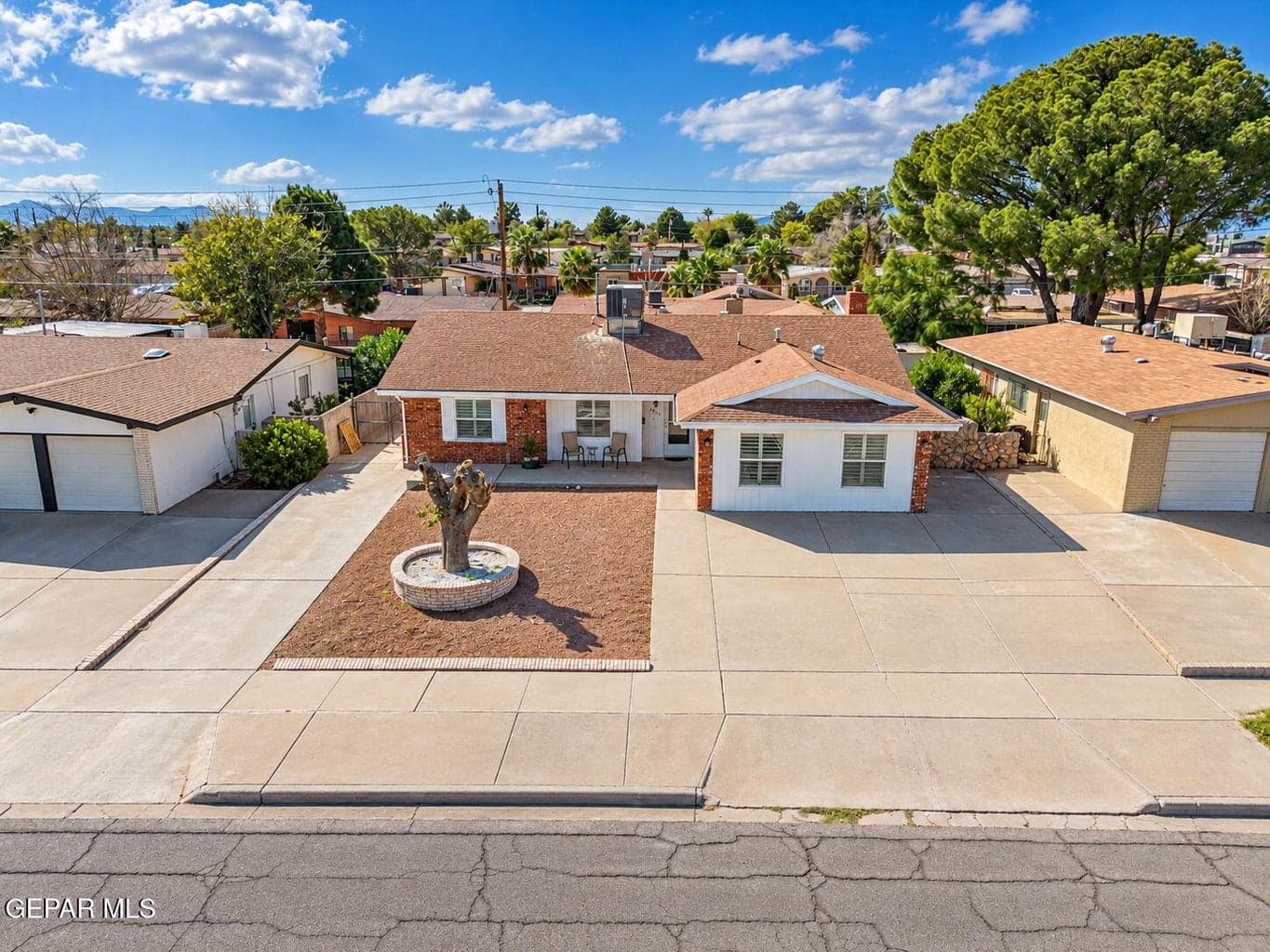 Single-story residence featuring a brick and stucco exterior, expansive concrete driveway, shingled roof, and a stone-rimmed landscape planter