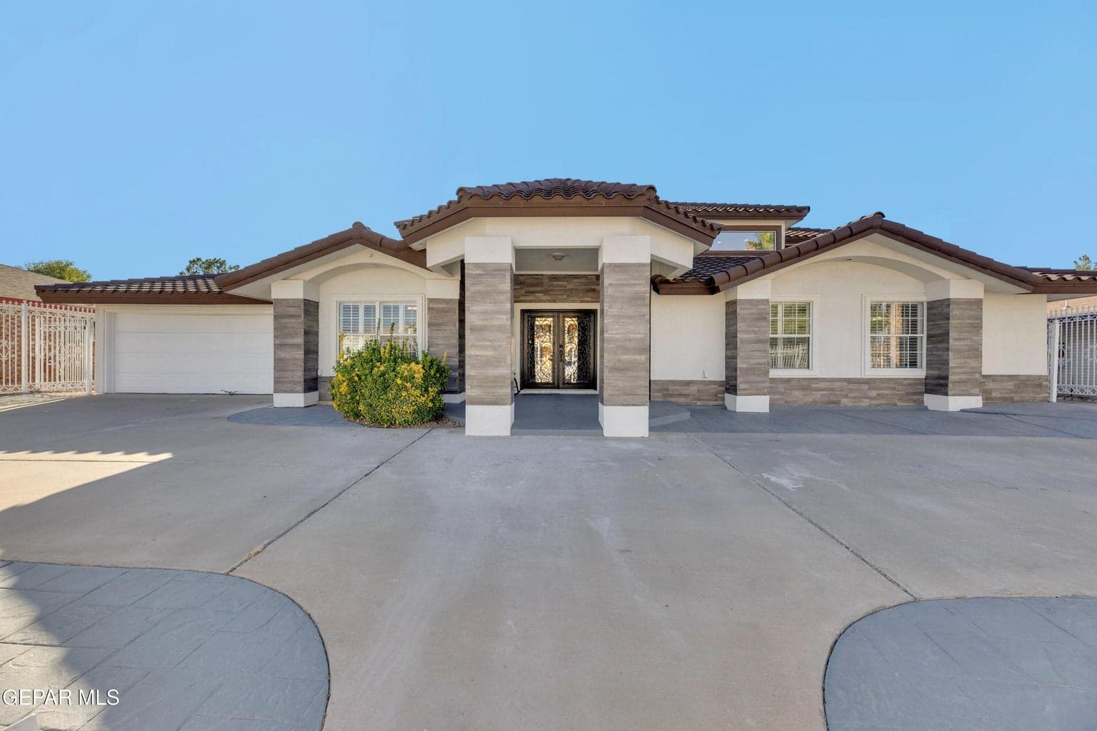 Single-story stucco exterior featuring tile roofing, stone-accented support columns, a recessed entryway with double doors, and a spacious concrete driveway